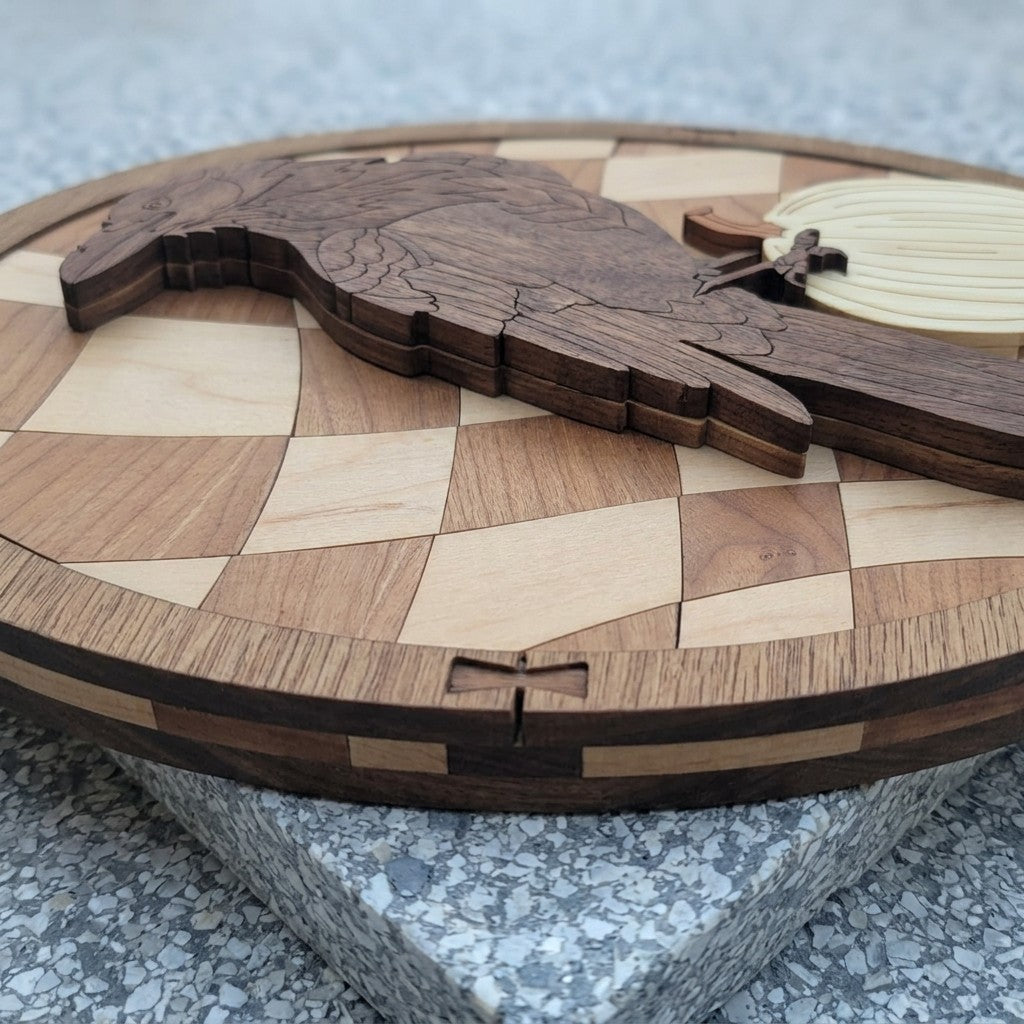 Side view of the oval’s layered edge, with stacked wood layers, a bowtie key inlay and the raised raven, resting on speckled stone.