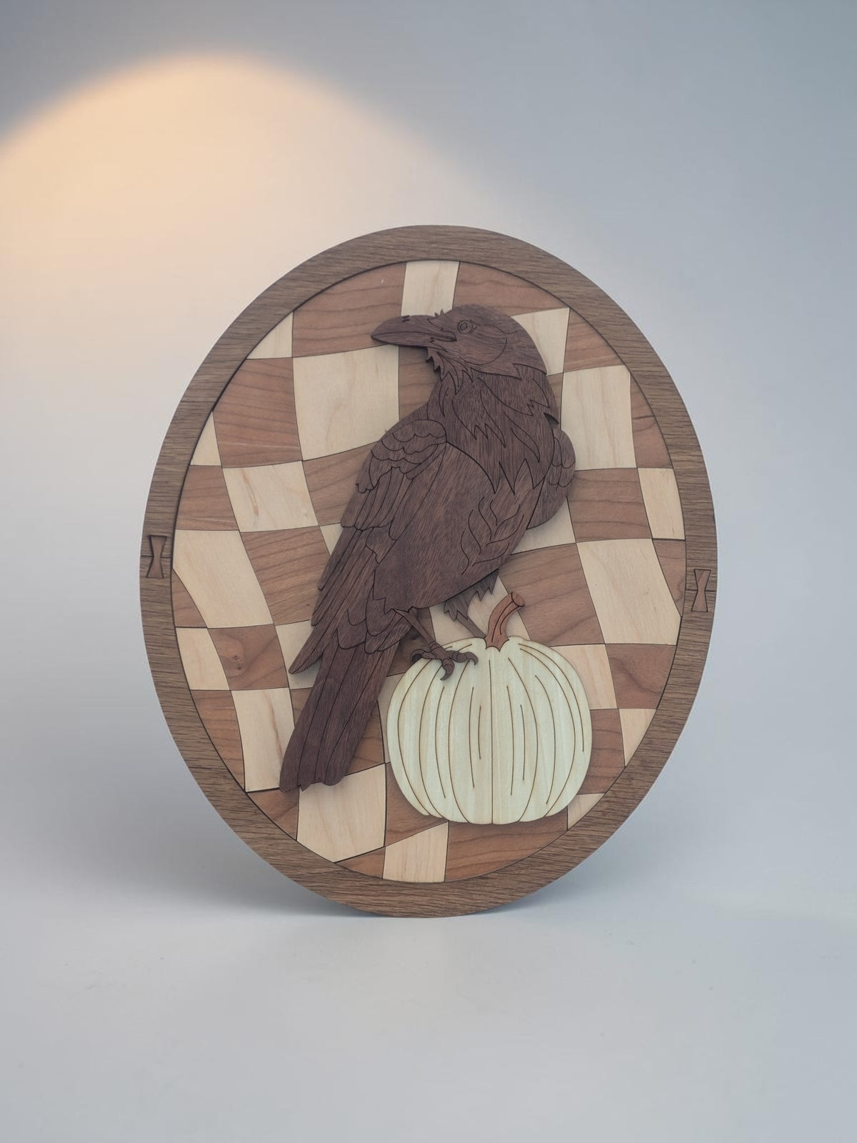 Raven and pumpkin wood wall art, a warped checkerboard oval in a walnut frame, standing against a soft gray backdrop with a warm glow.