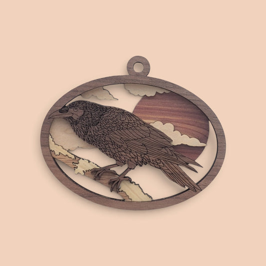 Raven wood ornament on a soft peach background, with the oval frame, hanging loop, sun, clouds and perched raven.