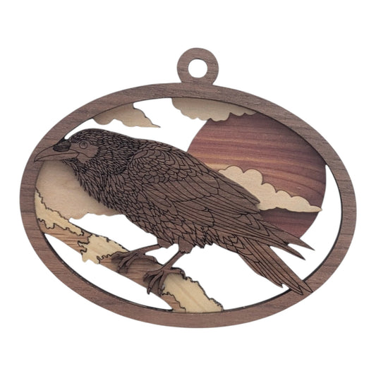 Layered wood Raven ornament, about 4 inches, with a raven perched on a branch in front of a large sun and clouds, in an oval frame.