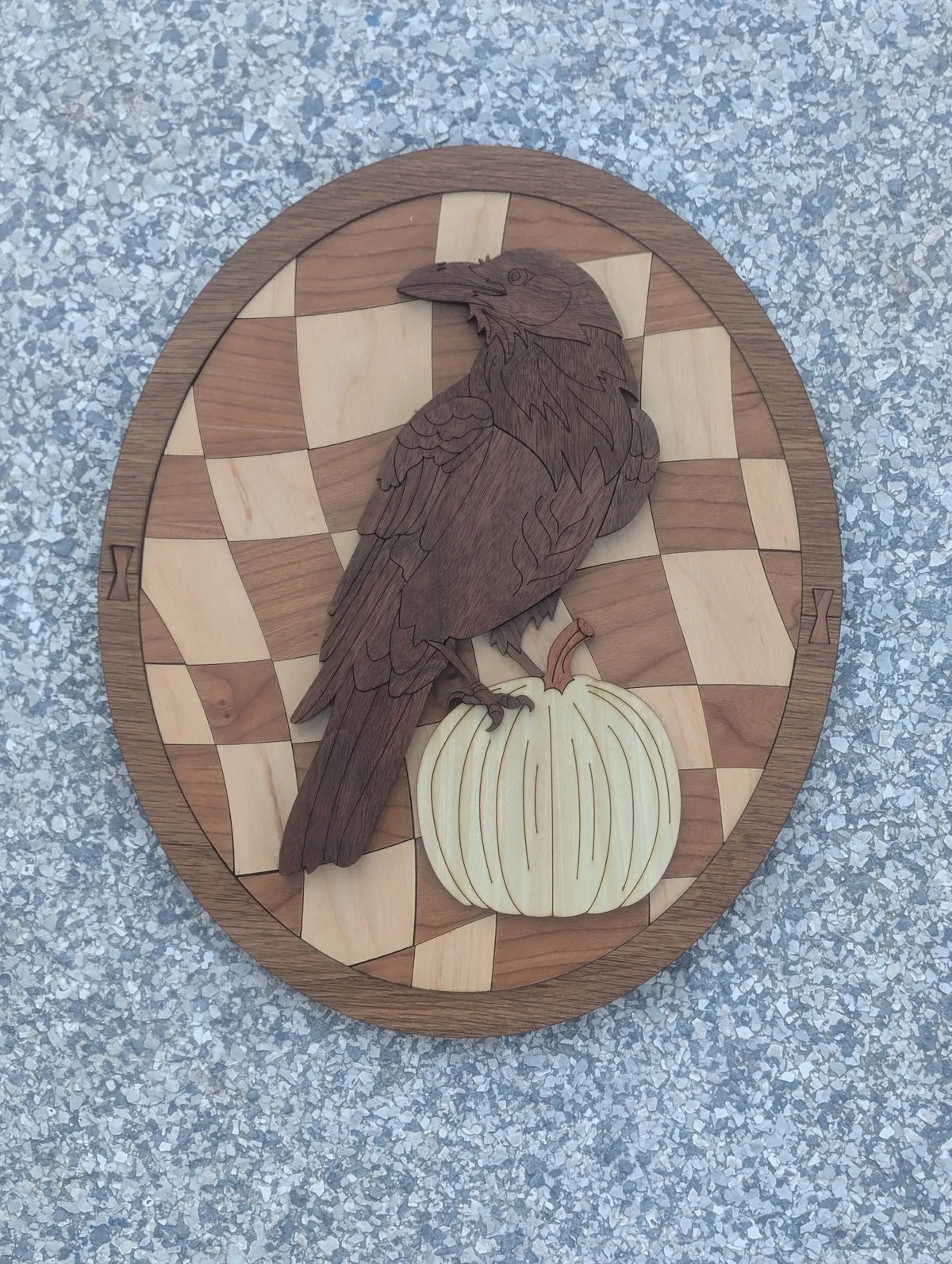 Handcrafted wooden wall plaque featuring a dark raven perched on a light pumpkin against a checkered wood background.